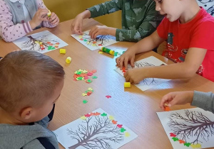 Dzieci przy stoliku dekorują listkami jesienne drzewko.